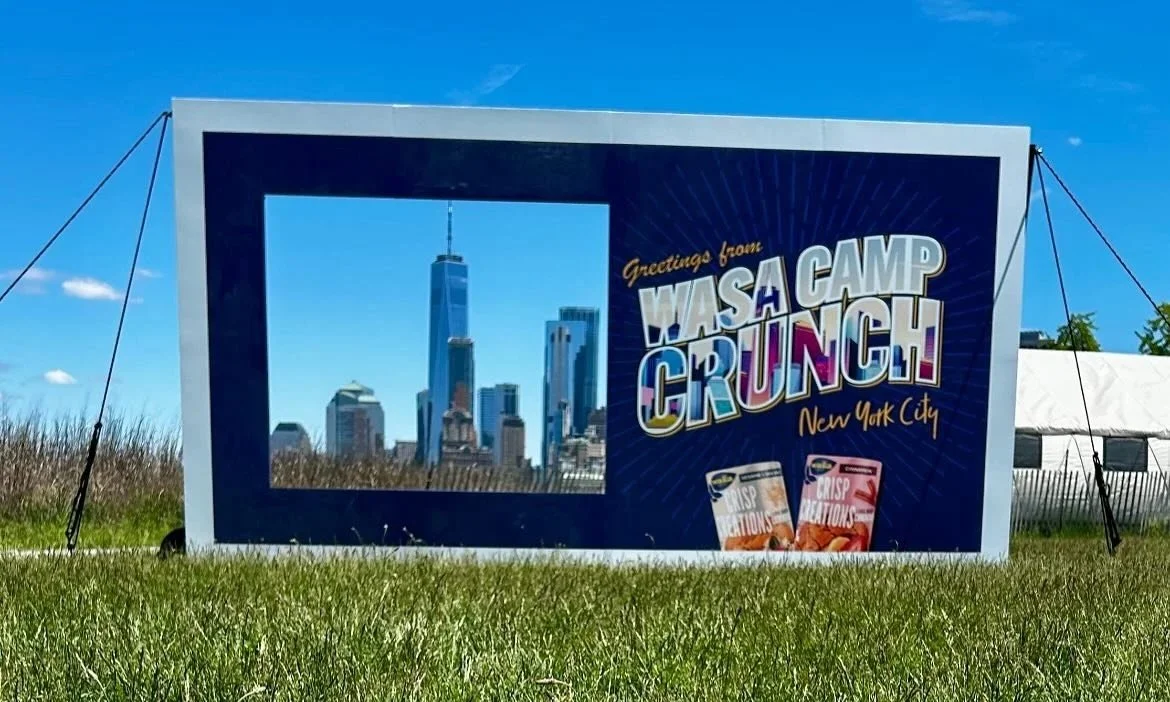 A large outdoor sign with a New York City skyline photo, and the text "Greetings from Wasa Camp Crunch! New York City" with images of snack products.