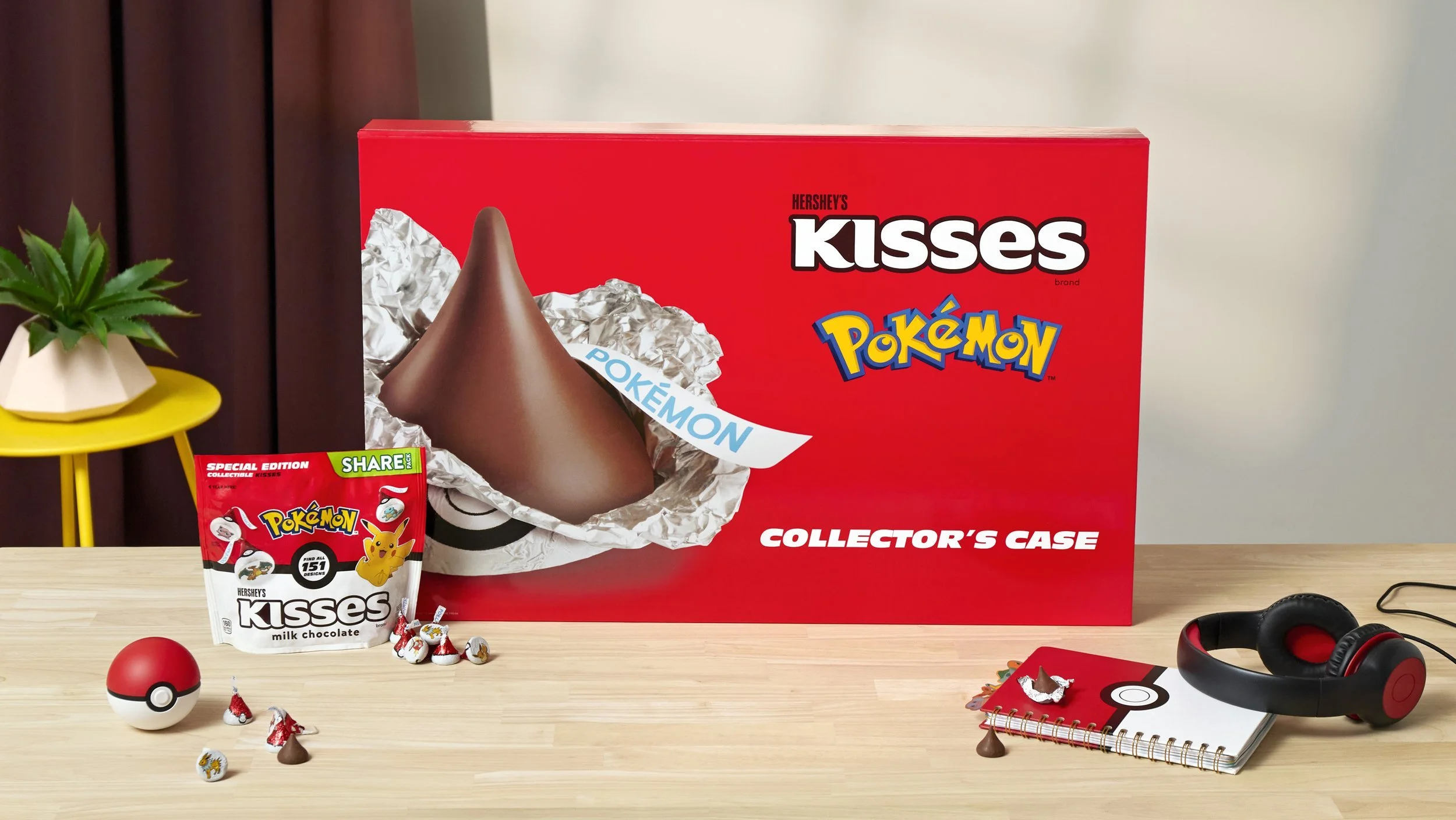 The image shows a Pokémon-themed collector's case box for Hershey's Kisses chocolates, with a Pikachu figurine and a Pokémon-themed small package. There are Pokémon candies, a Pokéball, and Pokémon-themed accessories like headphones and a notebook on a wooden table, with a small yellow table and a potted plant on the left side.