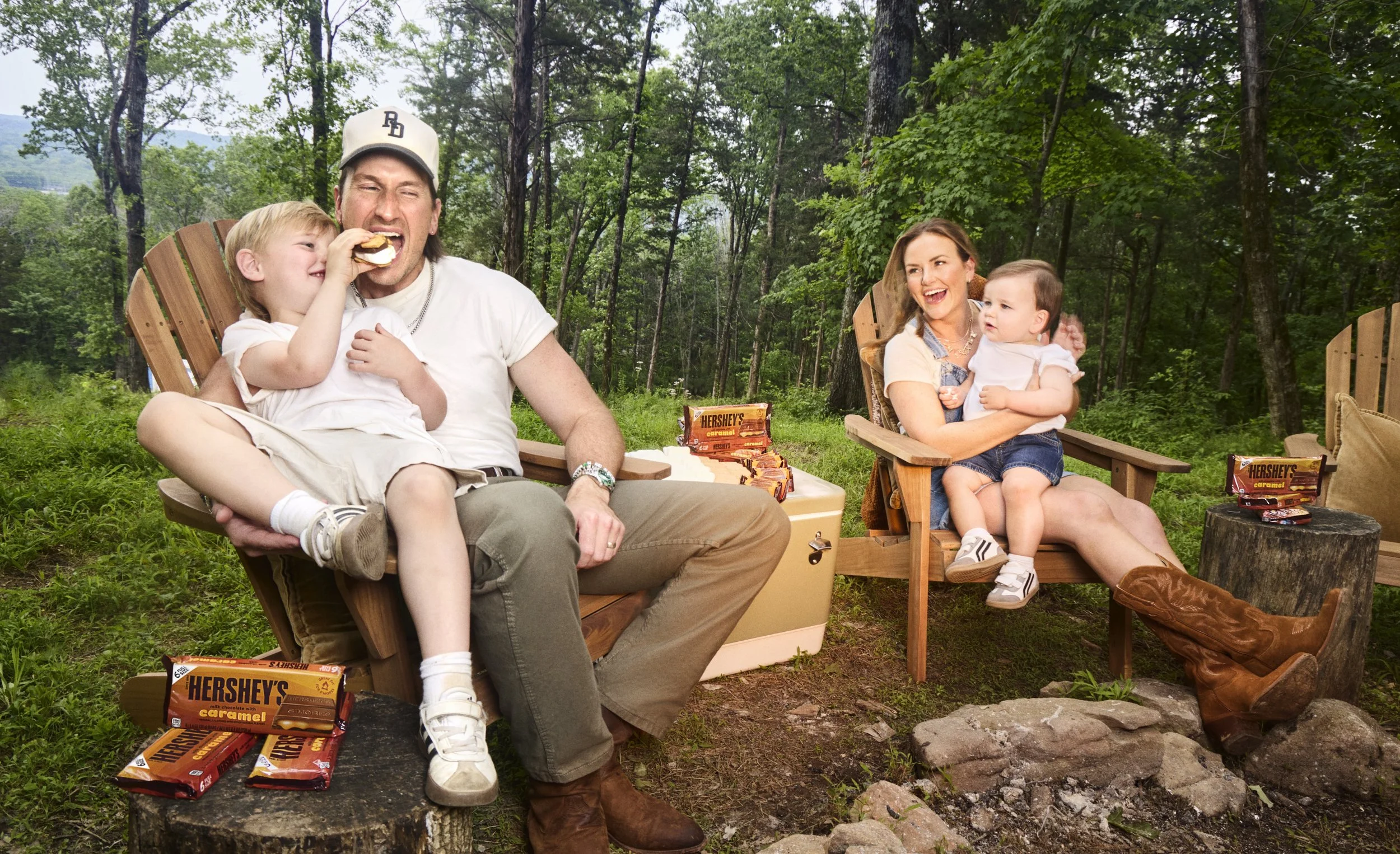A family of four enjoying an outdoor picnic in a wooded area, with chocolate bar snacks visible on the table and ground.