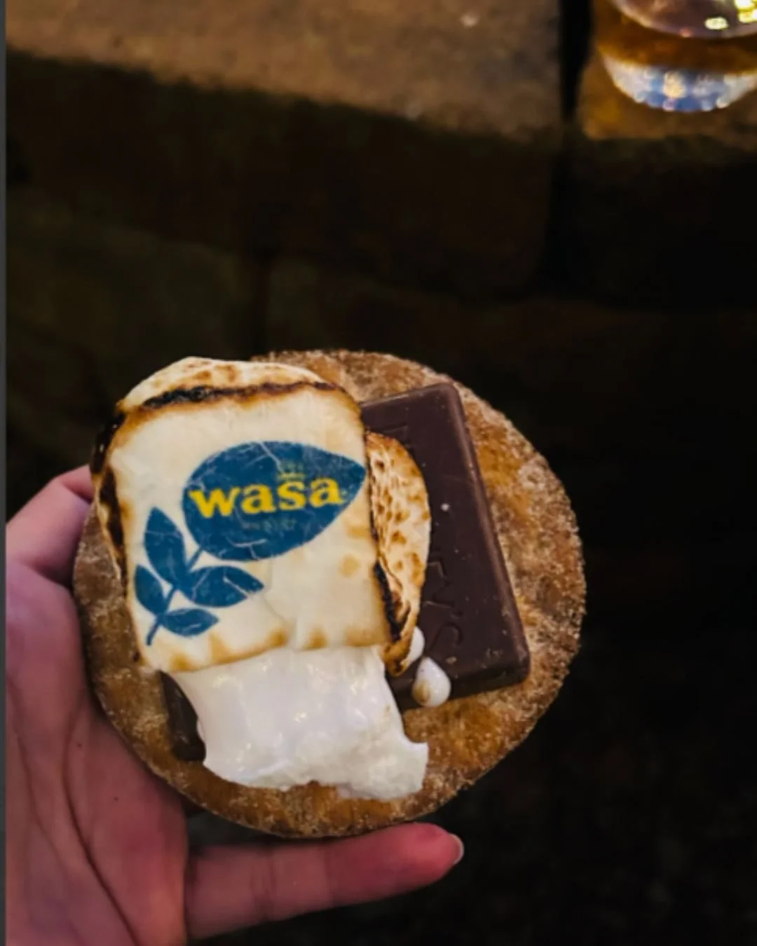 A person holding a round biscuit topped with caramelized apple, a piece of chocolate, dollops of cream, and a marbled spoon with the 'Wasa' logo.