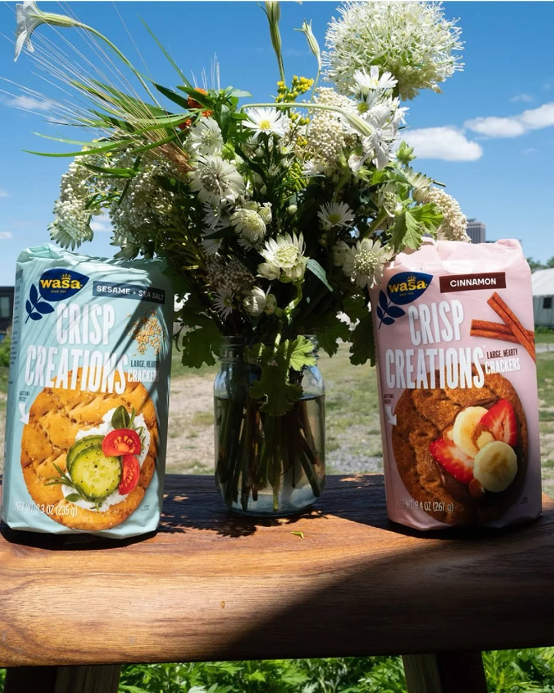 A bouquet of white flowers in a glass vase placed on a wooden table, with two bags of Wasa Crisp Creations crackers, one sesame sea salt flavor on the left and one cinnamon flavor on the right, against a backdrop of a blue sky with clouds.