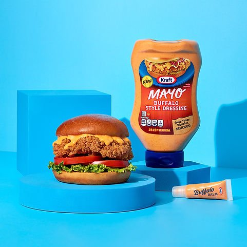 A burger with lettuce, tomato, cheese, and a sausage patty on a bun, placed on a blue circular platform. Behind it, there is a bottle of Kraft Mayo Buffalo Style Dressing and a small tube of Buffalo Balm, all set against a light blue background.