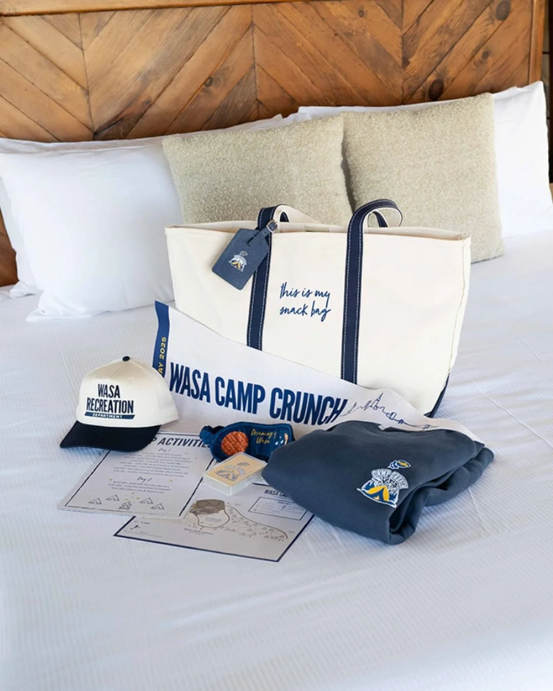 Collection of WASA River Recreation camping gear on a bed, including a white tote bag with navy handles and text, a matching cap, a gray t-shirt with a logo, a colorful laminated activity sheet, a small notepad, and other items, with a wooden headboard in the background.