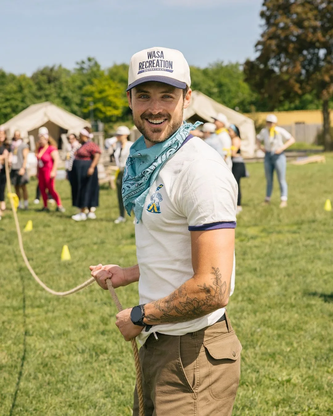 Dylan Efron holding a rope in a park during a community event with tents and people in the background.