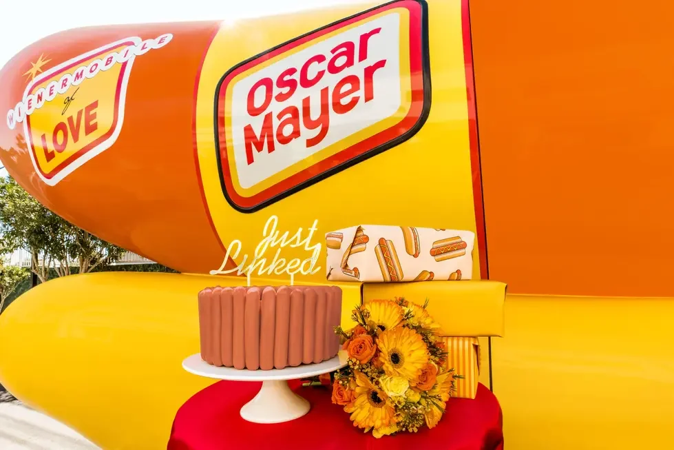 A large Oscar Mayer hot dog balloon with the words 'Wienermobile of Love' and 'Just Launched.' In front, there is a hot dog-shaped cake, a bouquet of flowers, and some gift boxes on a table.