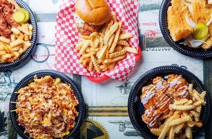 Four black takeout containers filled with food, including fries, fried chicken, and barbecue ribs, with a burger and fries on a red checkered paper in the center, all on a table.