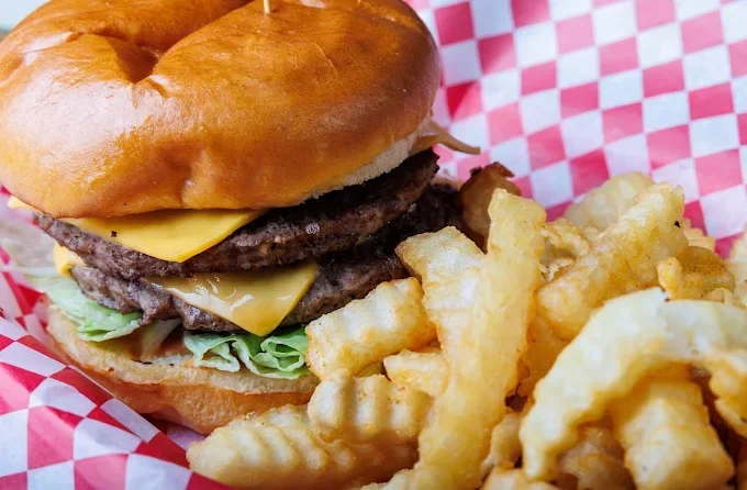 Hamburger with two beef patties, cheddar cheese, lettuce, and a bun, served with crinkle-cut French fries in a basket lined with red and white checkered paper.