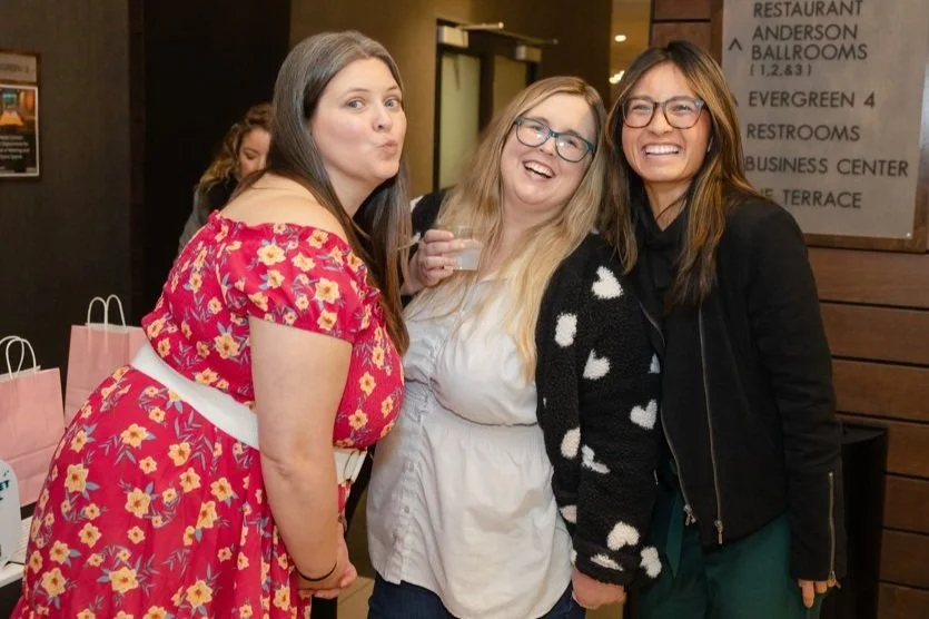 Three women smiling and posing together indoors at a women's small business expo, with pink swag bags on a table nearby.