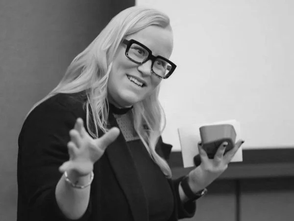 A woman with long blonde hair wearing glasses and a dark top, smiling, and holding a smartphone in her right hand while gesturing with her left hand. She is presenting on neurodivergent tips for female business owners.