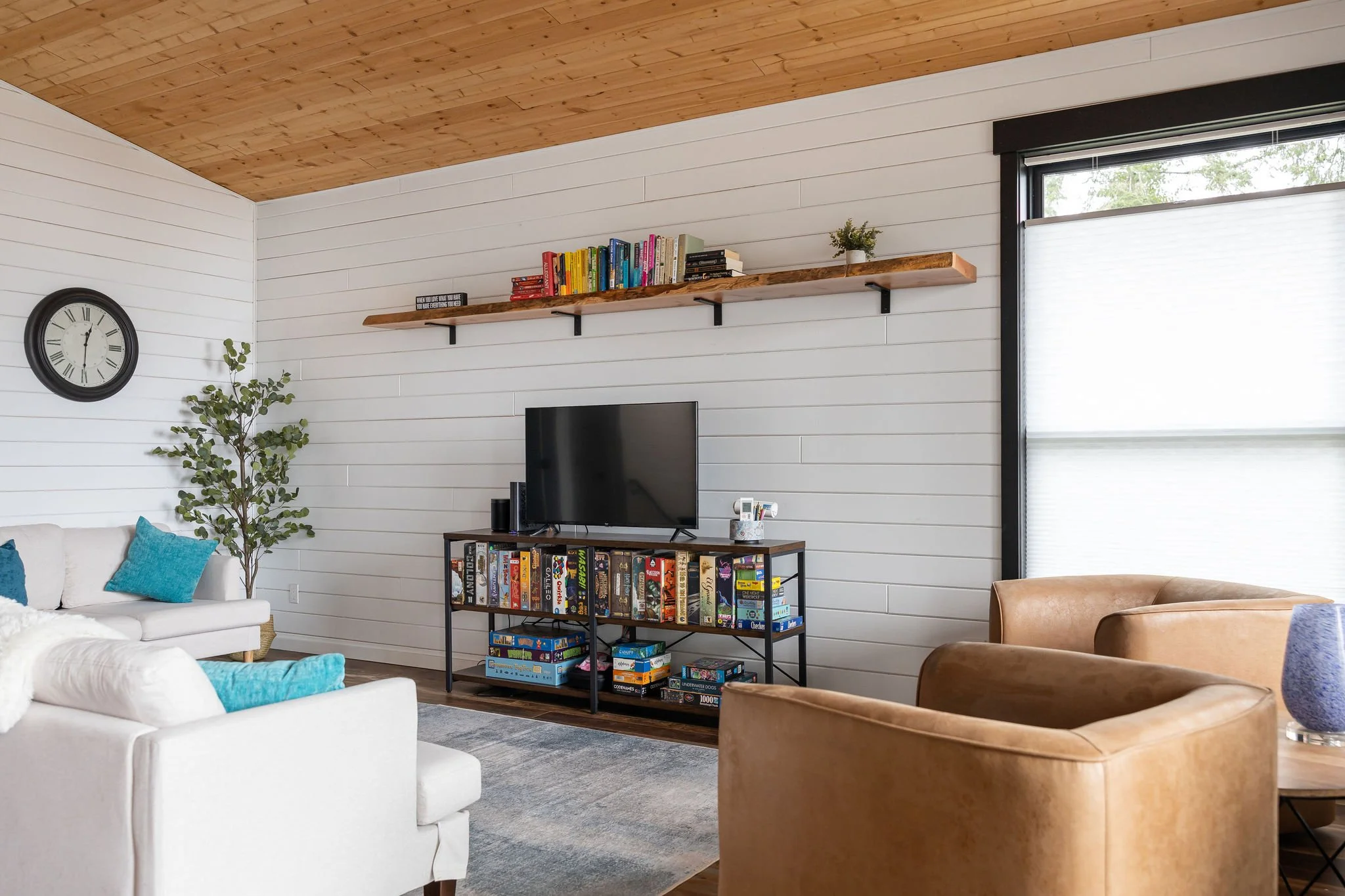 Living room with white shiplap walls, a wooden ceiling, a black wall clock, a large window with blinds, a potted plant, a white sofa with teal pillows, a TV on a black and wood media stand filled with board games, a brown armchair, and a coffee table.