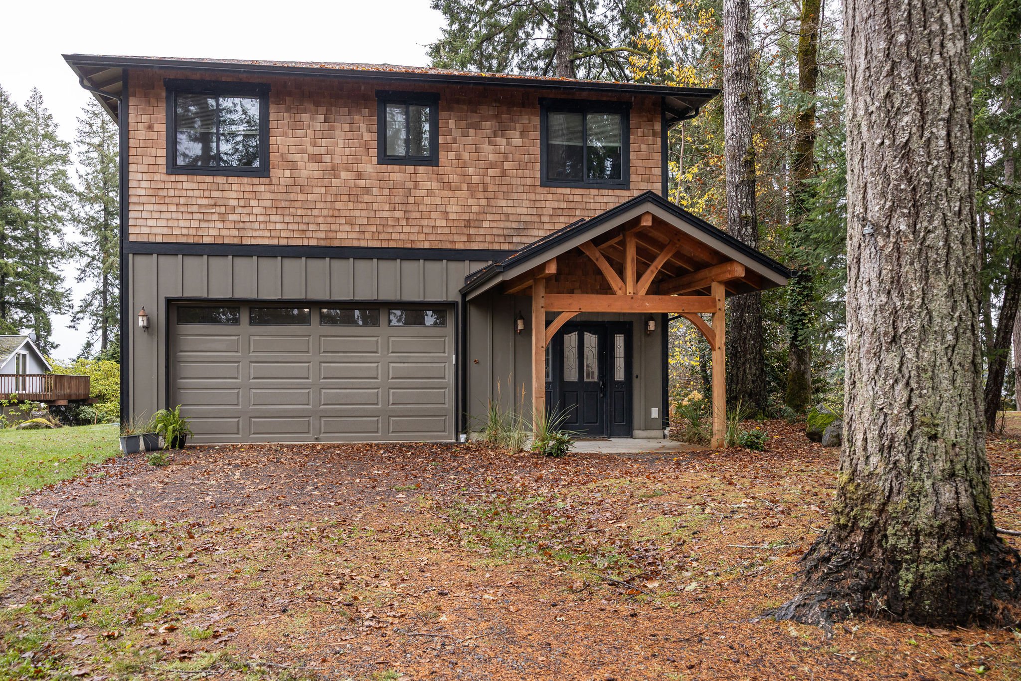Front view of a two-story house with a gray garage door, wooden shingles on the upper floor, and a covered entryway with wood beams, set among tall trees with leaves on the ground.