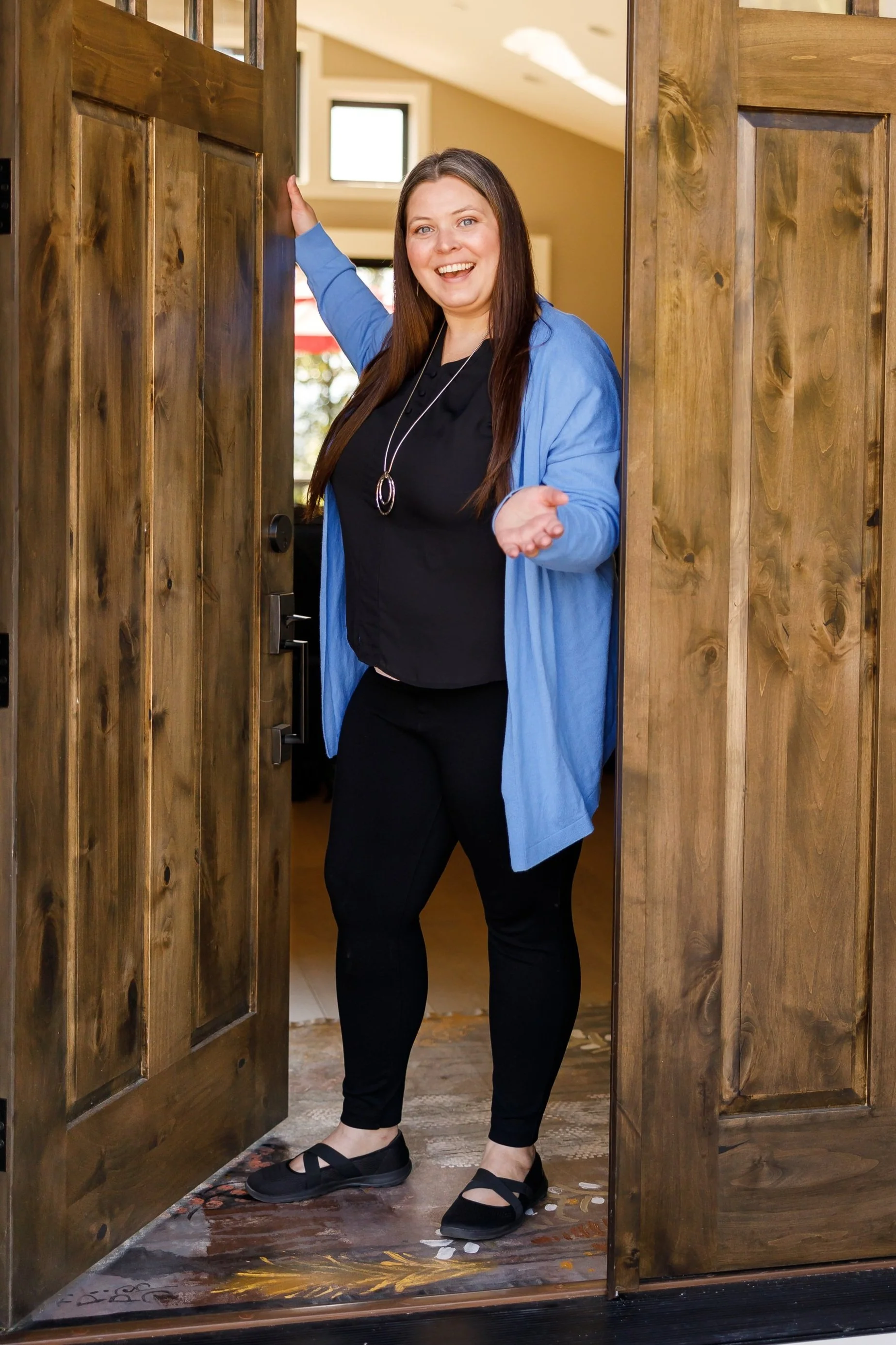 A cheerful woman standing at an open wooden door, indoors with windows overhead, wearing black pants, black shoes, a black top, and a blue cardigan, smiling and gesturing with her hand.
