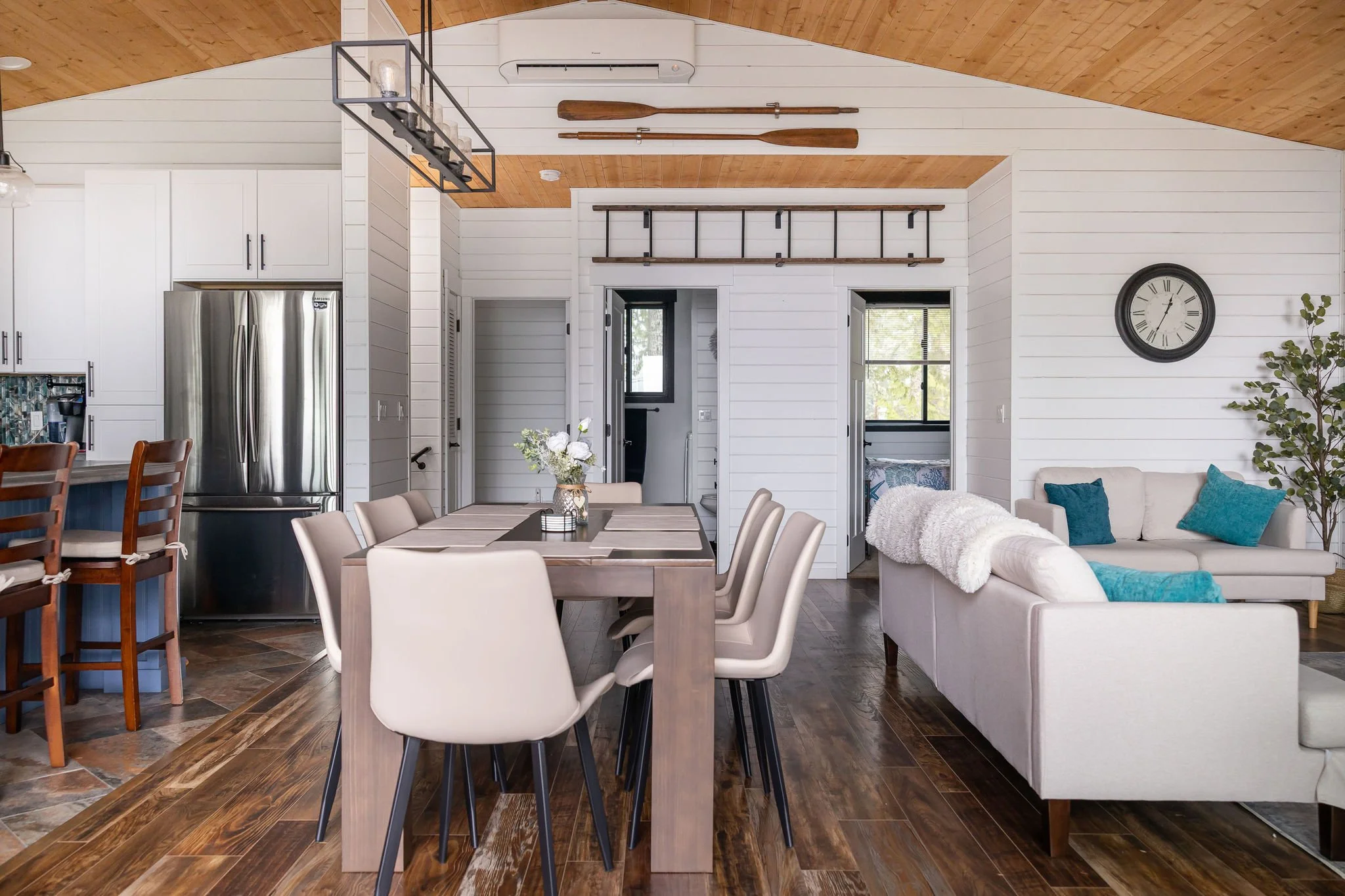 Open-concept living and dining area in a modern home with white shiplap walls, wooden flooring, a white couch with blue pillows, a dining table with beige chairs, a wall clock, and decorative paddles on the wall.