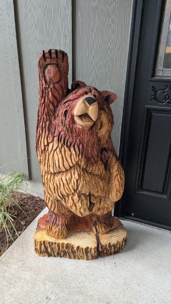 Wooden carved bear standing on a tree stump with one arm raised, positioned outside next to a house door.