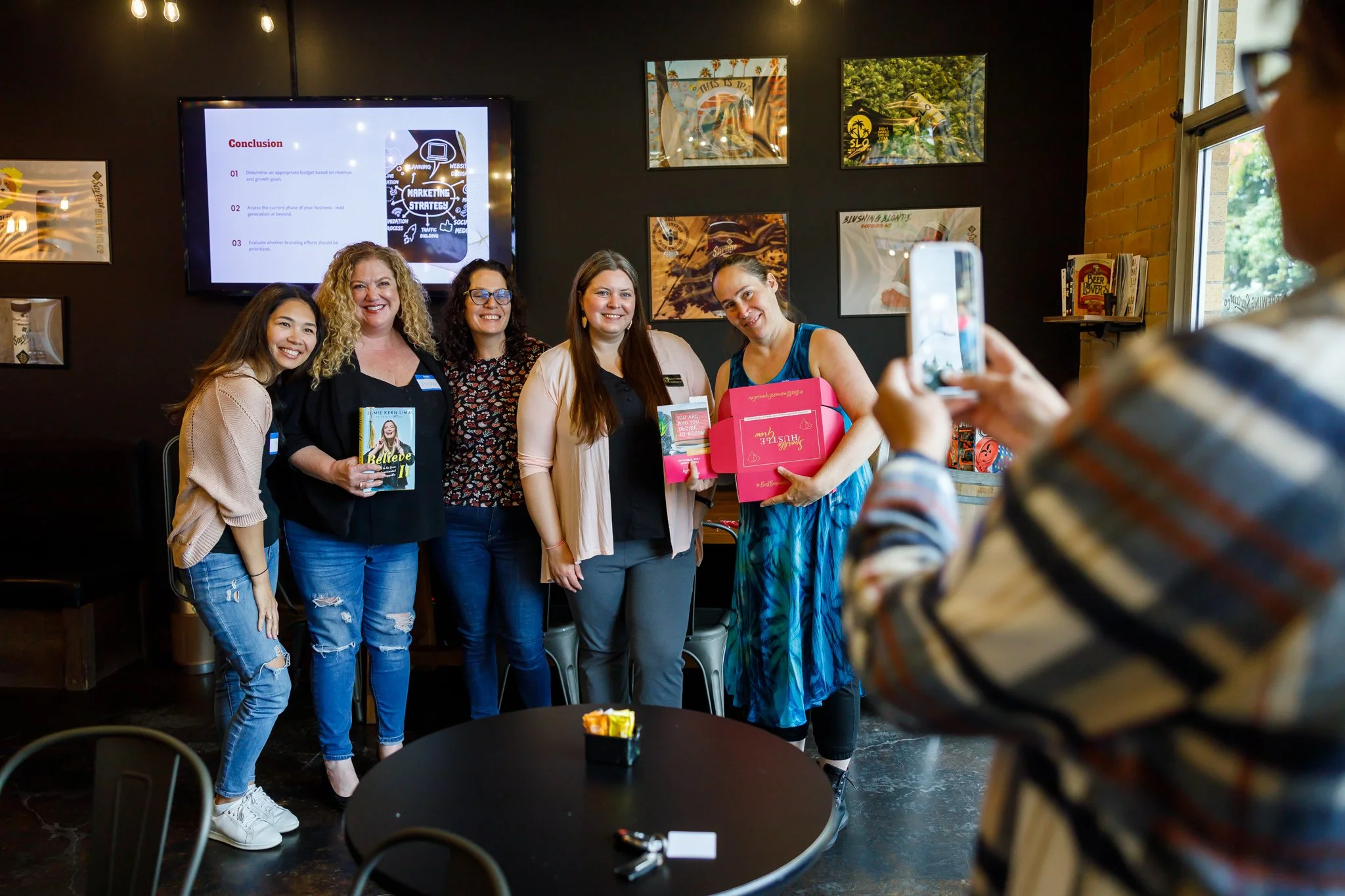 Group of five women posing for a photo in a cafe, one woman holding a book, another holding a pink box, while a woman in a striped shirt takes a picture with her phone. A screen with presentation slides is visible in the background.