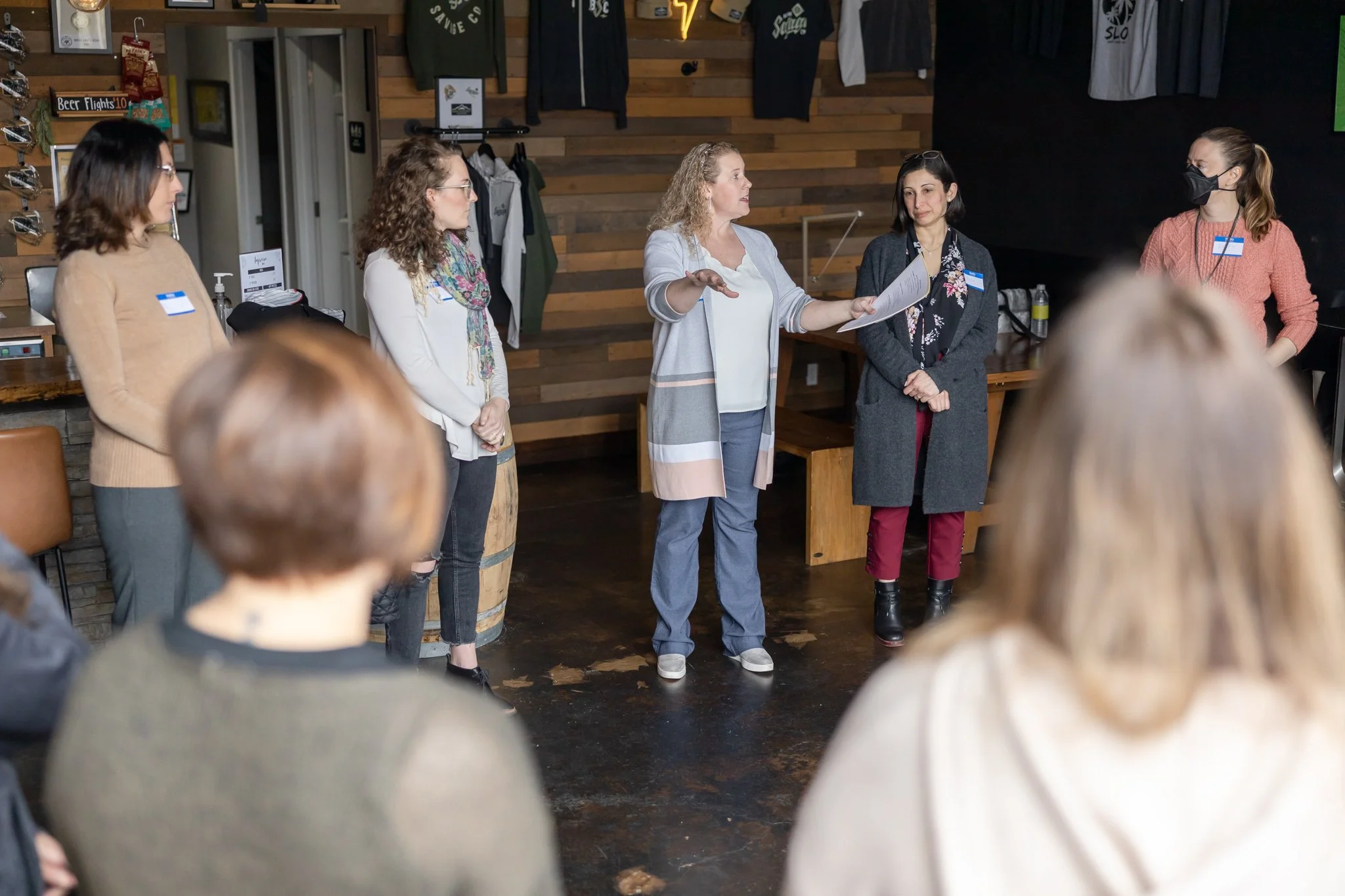 A woman in a light gray cardigan appears to be speaking to a group of women in a casual indoor setting, possibly a cafe or meeting room, with wood-paneled walls and hanging jackets.