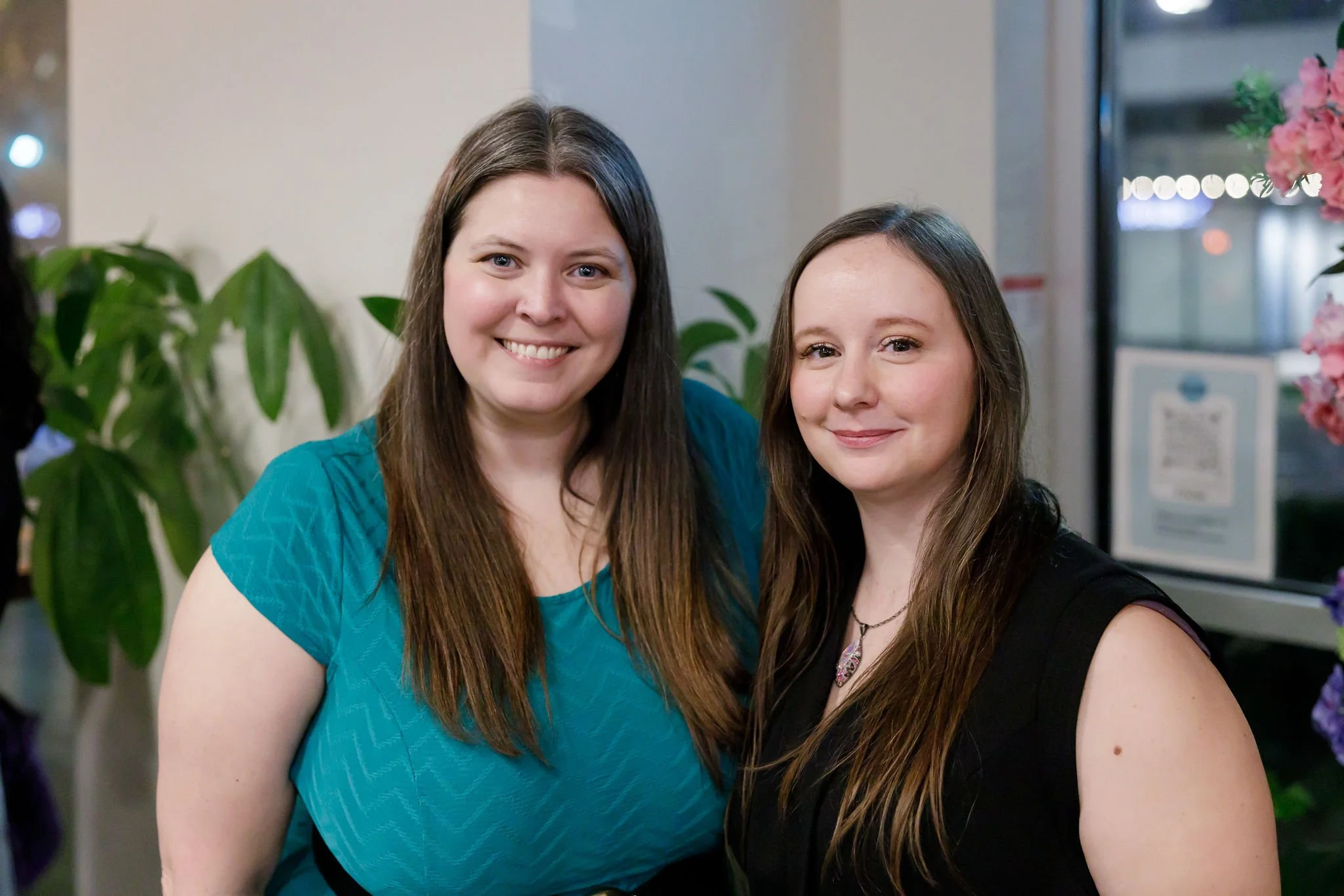 Two women smiling indoors, standing close together with plants and flowers in the background.