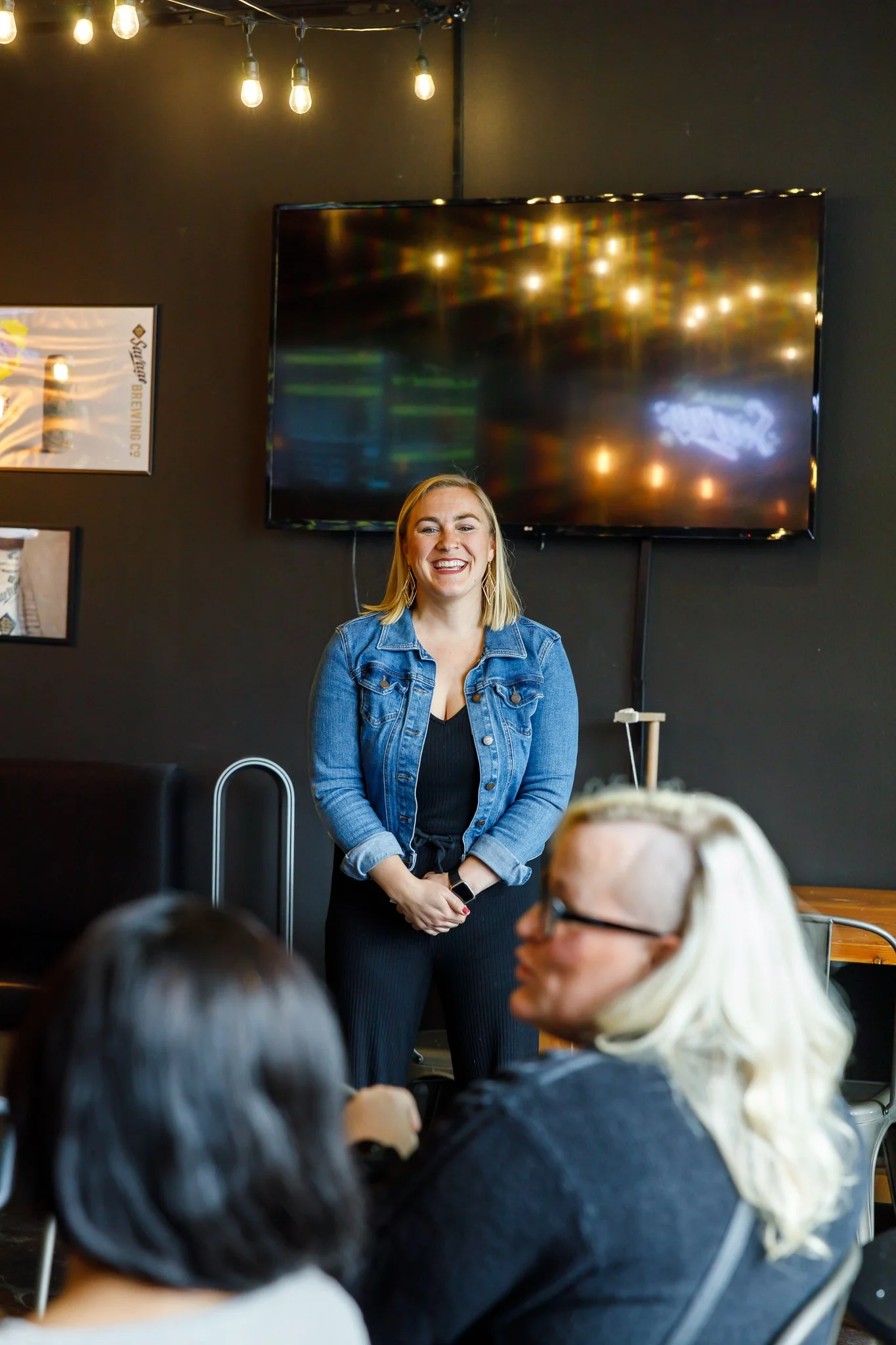 A woman smiling and standing in front of a black wall with a large television screen at a gathering or meeting, with people seated in front of her.