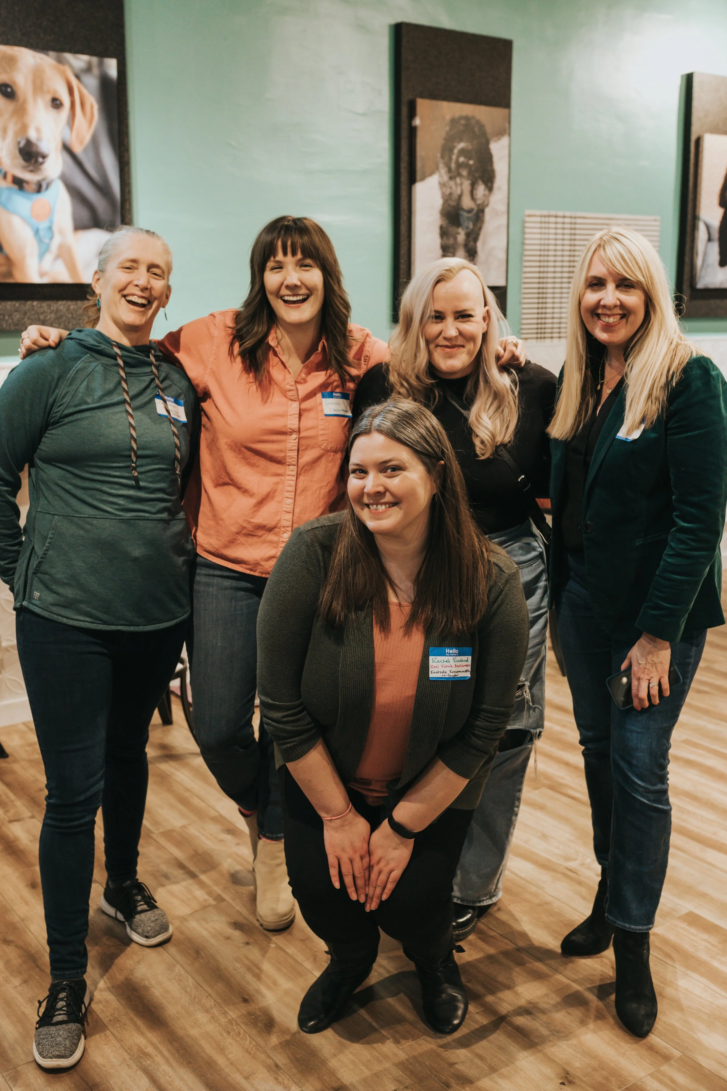 A group of five women smiling and posing together indoors, with dog portraits on the wall behind them.