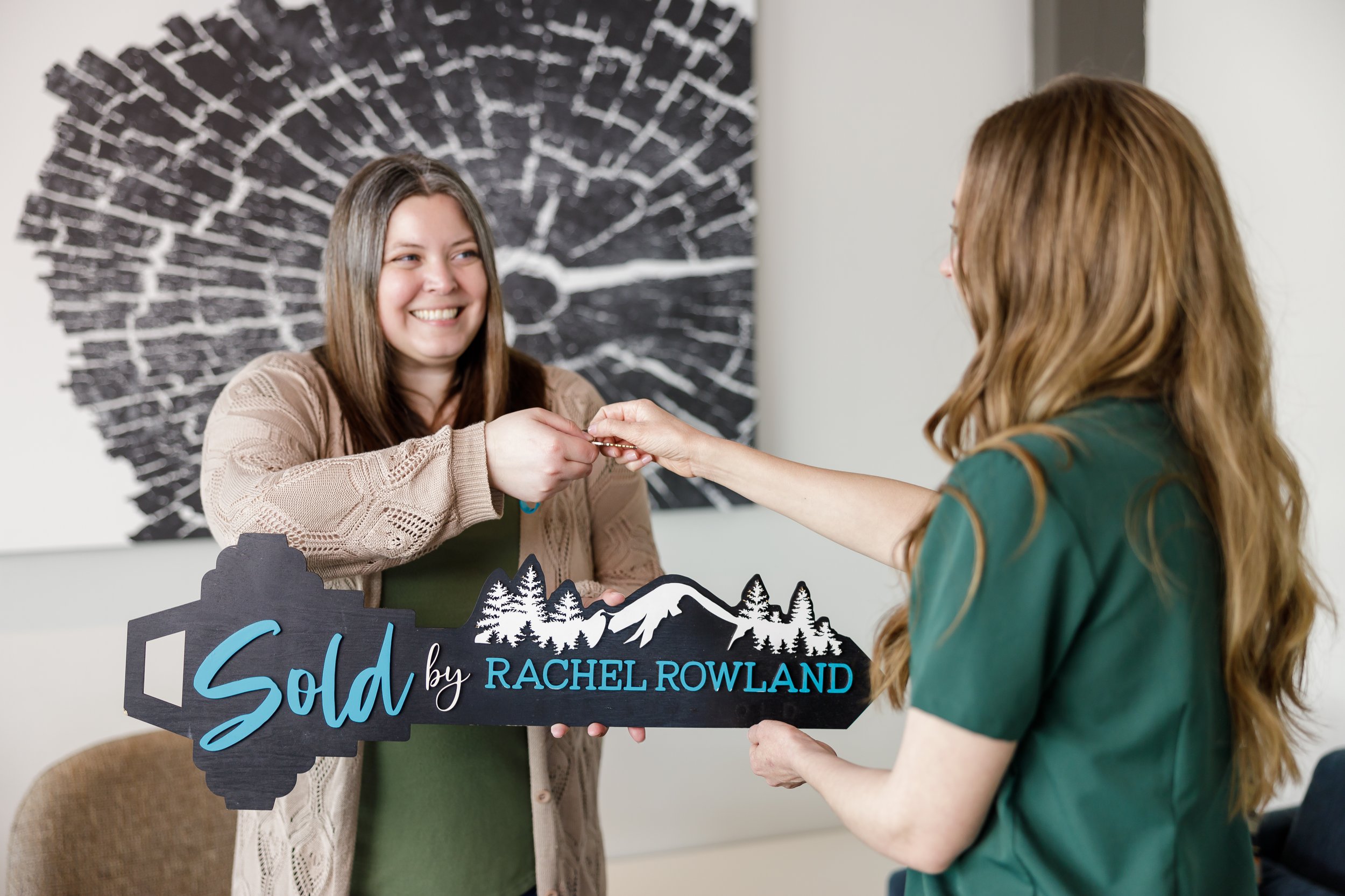 A woman receiving a key from another woman during a home sale, with a key-shaped sign that reads "Sold by Rachel Rowland" with a mount rainier and trees design.