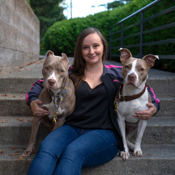 A woman sitting on outdoor stone steps holding two pit bull dogs, one on each side, with a green hedge and railing in the background. She is Nikki Johnson, owner of Pickles Playland and At Your Feet Dog Training.