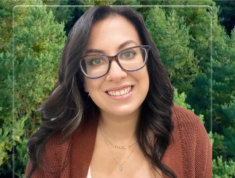 A woman with long dark wavy hair, glasses, and a nose ring smiling outdoors with green trees in the background.