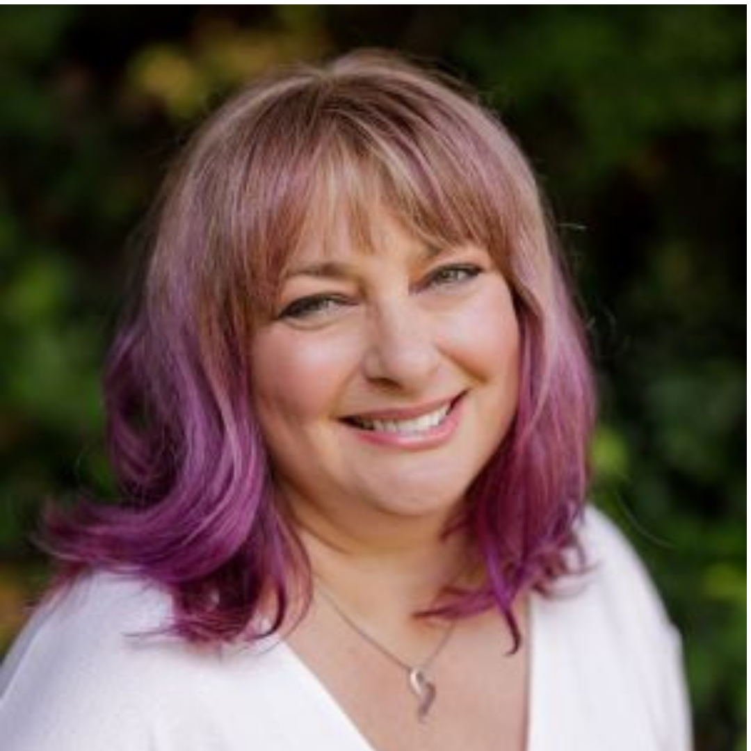 A woman with shoulder-length pink and purple hair, smiling outdoors with a blurred green background.