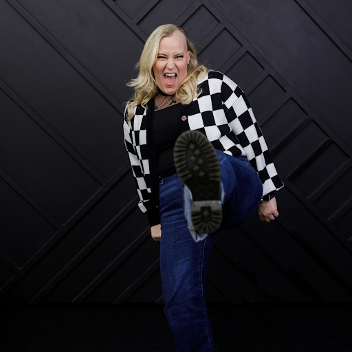 Woman with blonde hair making a playful face, lifting her foot toward the camera, wearing a black top, checkered black and white jacket, and blue jeans, against a dark geometric background.