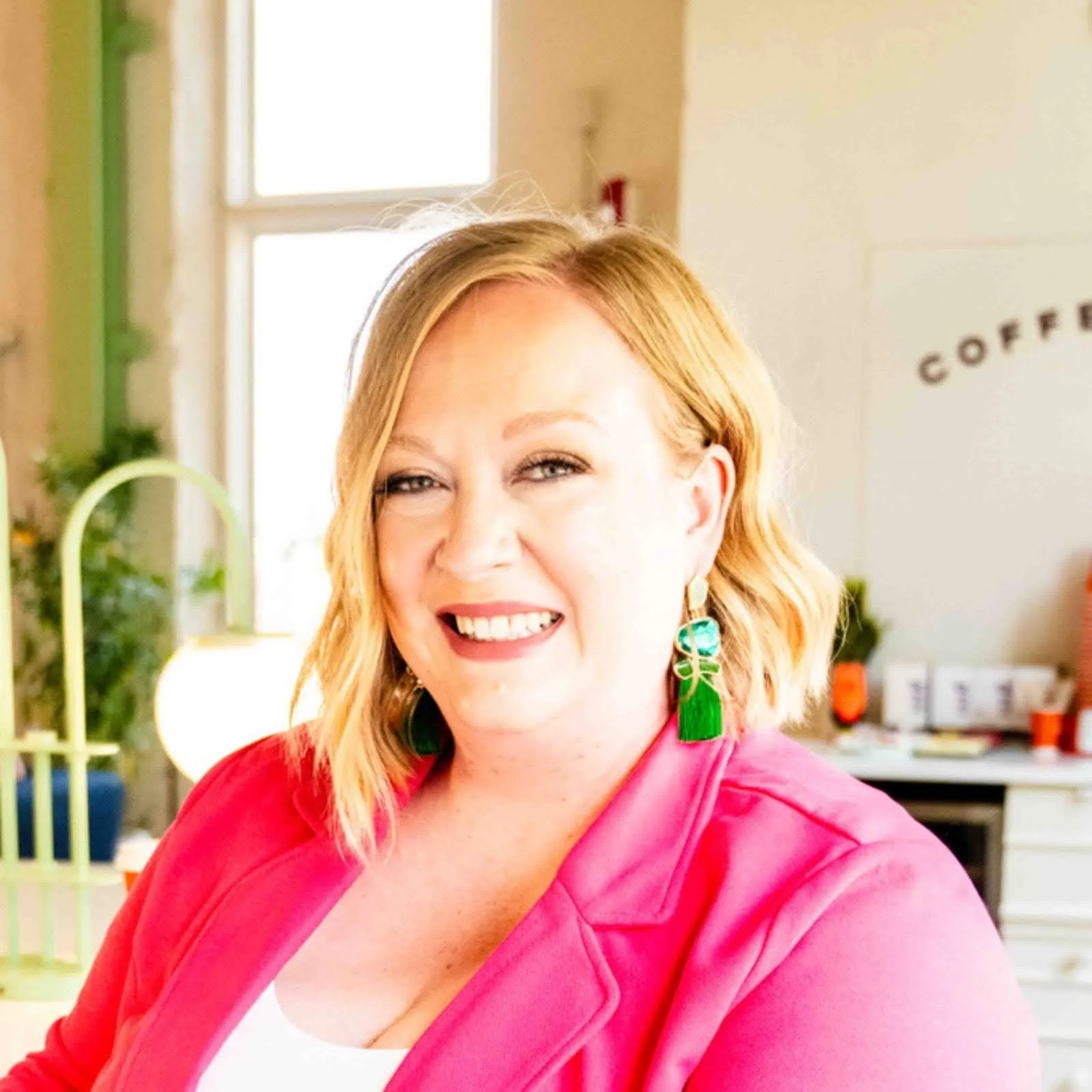 smiling woman with shoulder-length blonde hair, wearing a pink blazer and green earrings, in a bright room with sunlight coming through a window and a coffee sign in the background