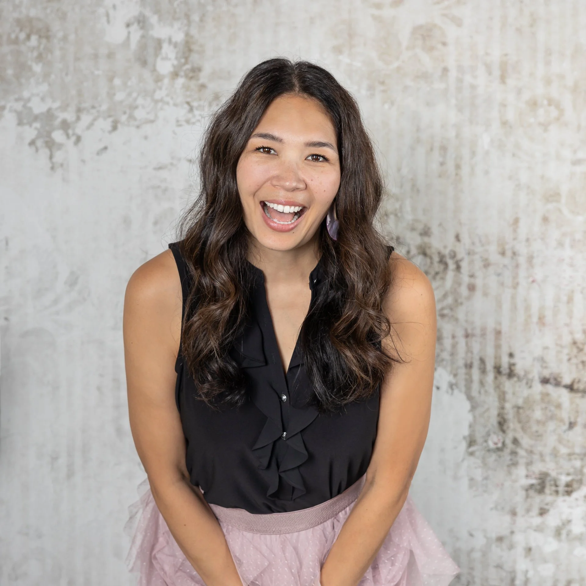 A woman with long, wavy dark hair and to earrings, wearing a black sleeveless top and a pink skirt, smiling against a textured wall background.