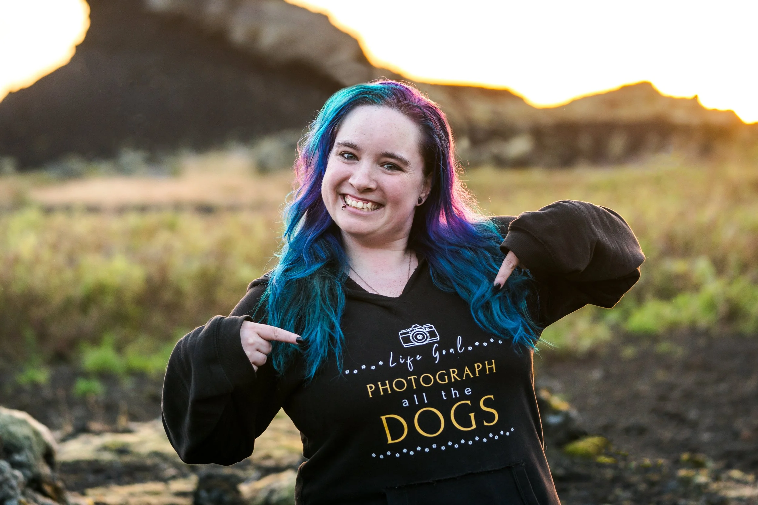 Young woman with multicolored blue and purple hair smiling outdoors during sunset, wearing a black hoodie that says "Life Goal... PHOTOGRAPH all the DOGS" and pointing to her hoodie.