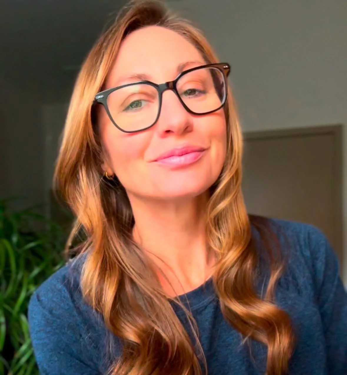 A woman with long, wavy brown hair and glasses smiling at the camera, in an indoor setting.