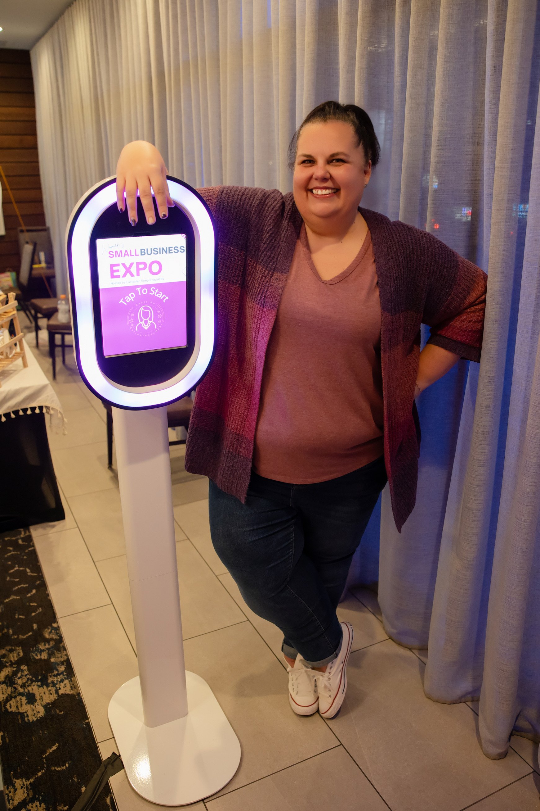 A woman standing indoors smiling next to an illuminated sign that reads 'Small Business Expo' and 'Tap To Start' with a graphic of a woman.