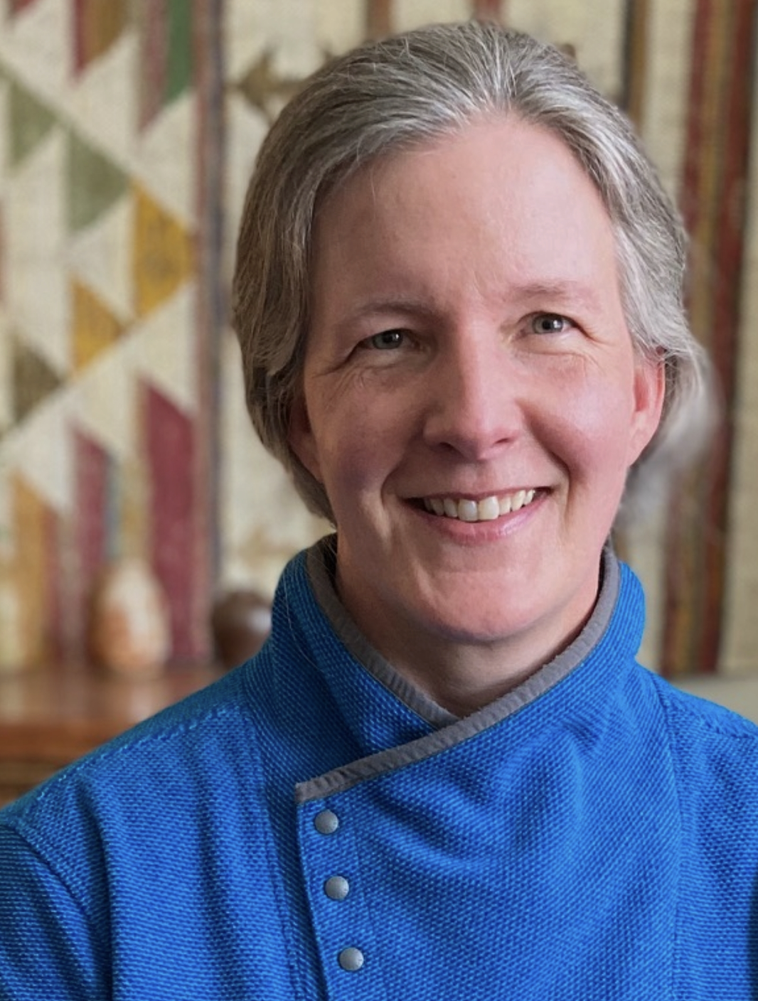 A middle-aged woman with short gray hair, wearing a blue chef's jacket, smiling in front of a patterned quilted wall hanging.