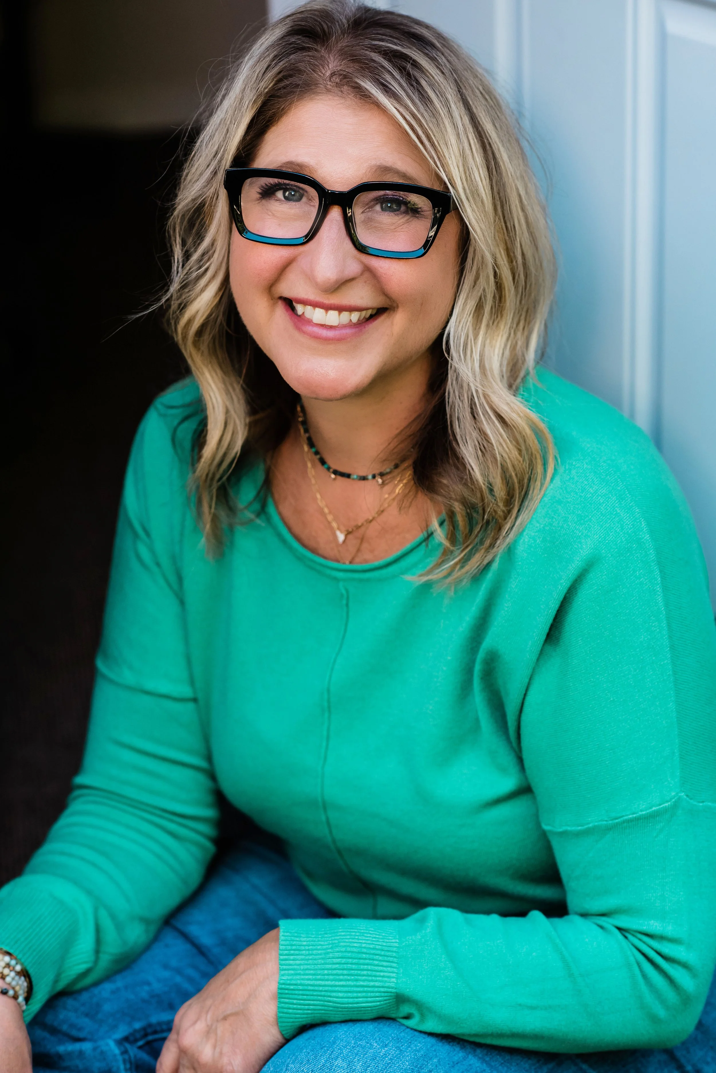 A smiling woman with shoulder-length blonde hair, wearing black glasses, a green sweater, and layered necklaces, sitting near a window.