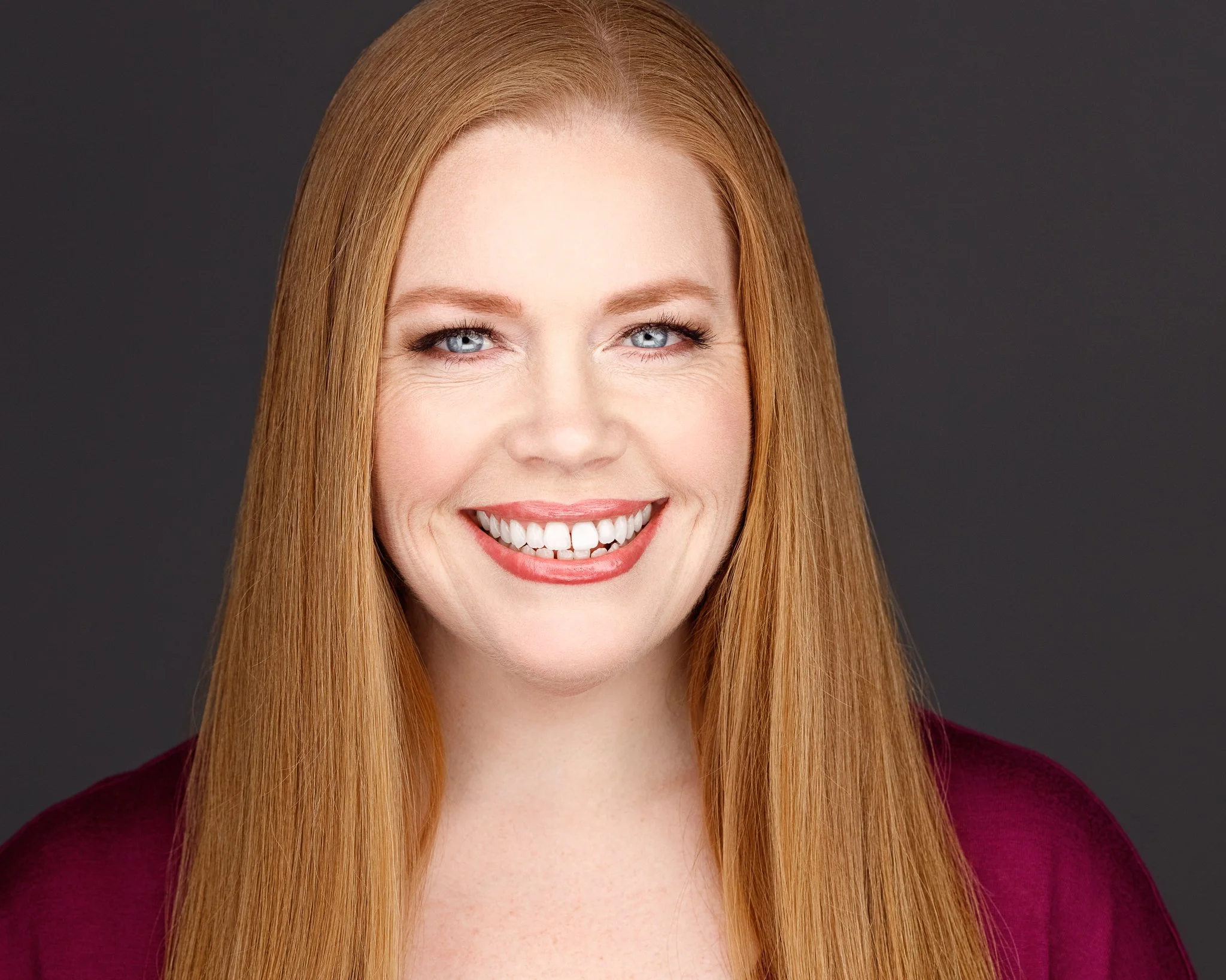 A woman with long red hair and blue eyes smiling, wearing a dark red top, against a plain background.