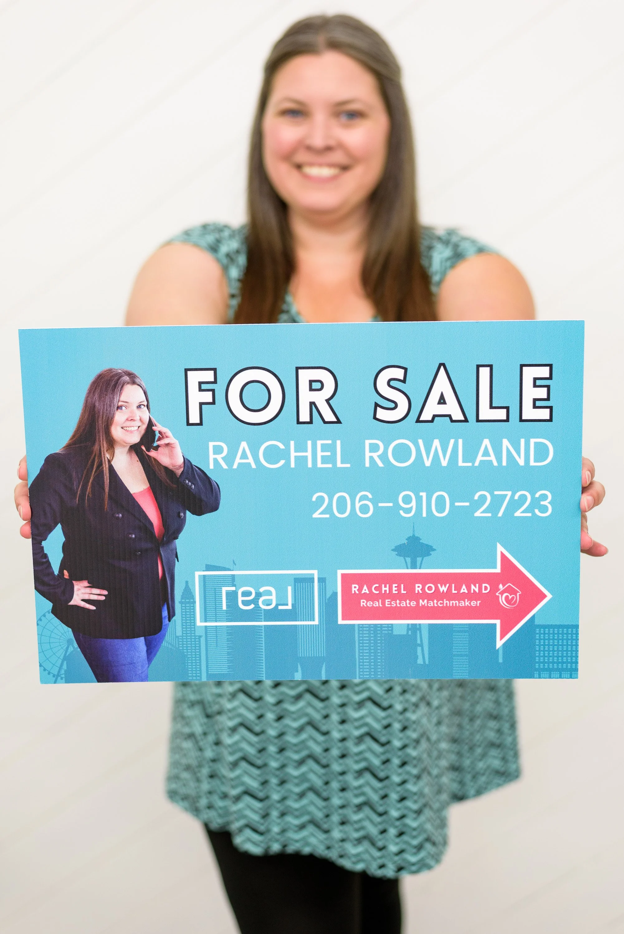 Rachel Rowland wearing a teal an black dress while  holding a for sale sign with her photos, phone number, real logo, and Seattle skyline in the background.