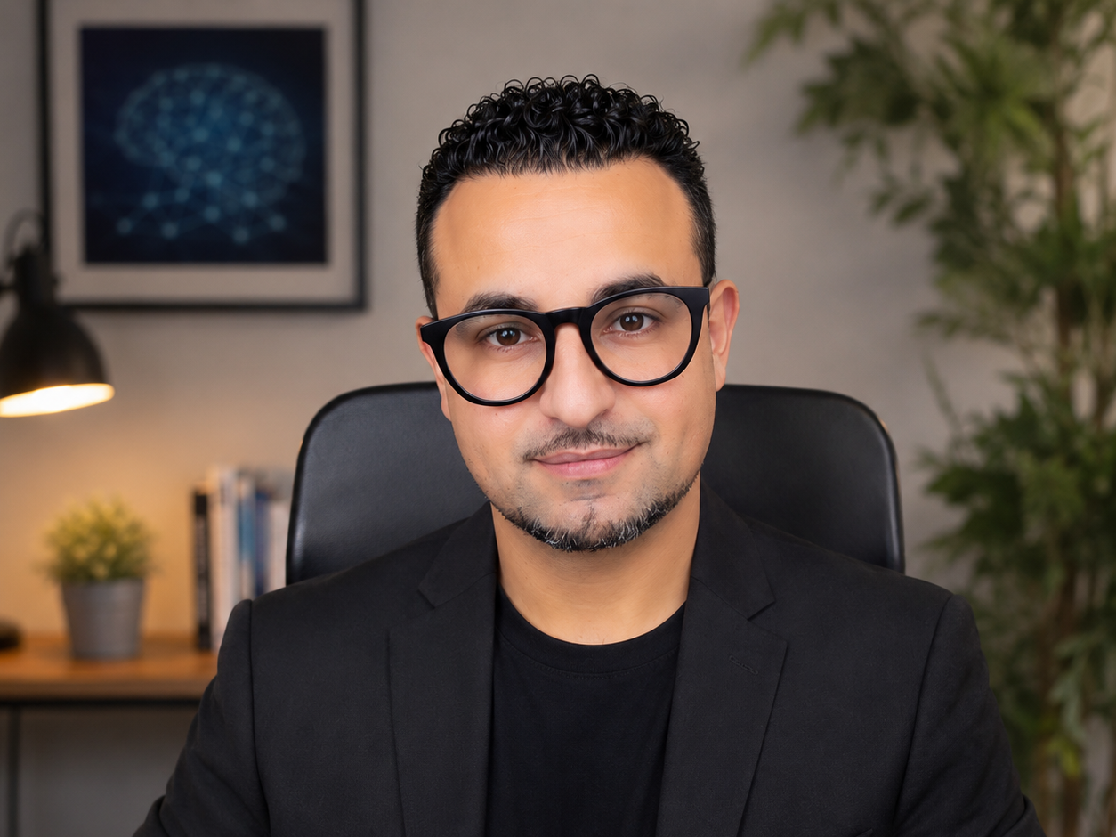 Un homme souriant, portant des lunettes noires, en costume noir, assis dans un bureau avec une plante verte en arrière-plan.