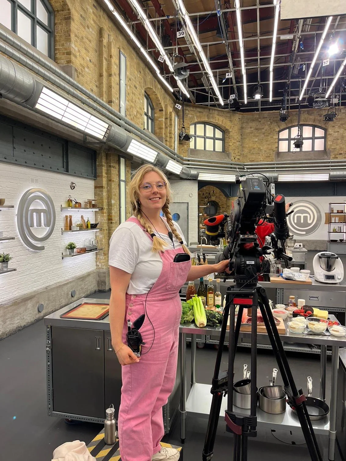 A woman in pink overalls and glasses stands in a cooking competition kitchen, smiling at the camera beside a professional camera on a tripod. The kitchen has a modern industrial look with exposed brick walls, large windows, and professional cooking equipment, with the MasterChef logo on the wall.