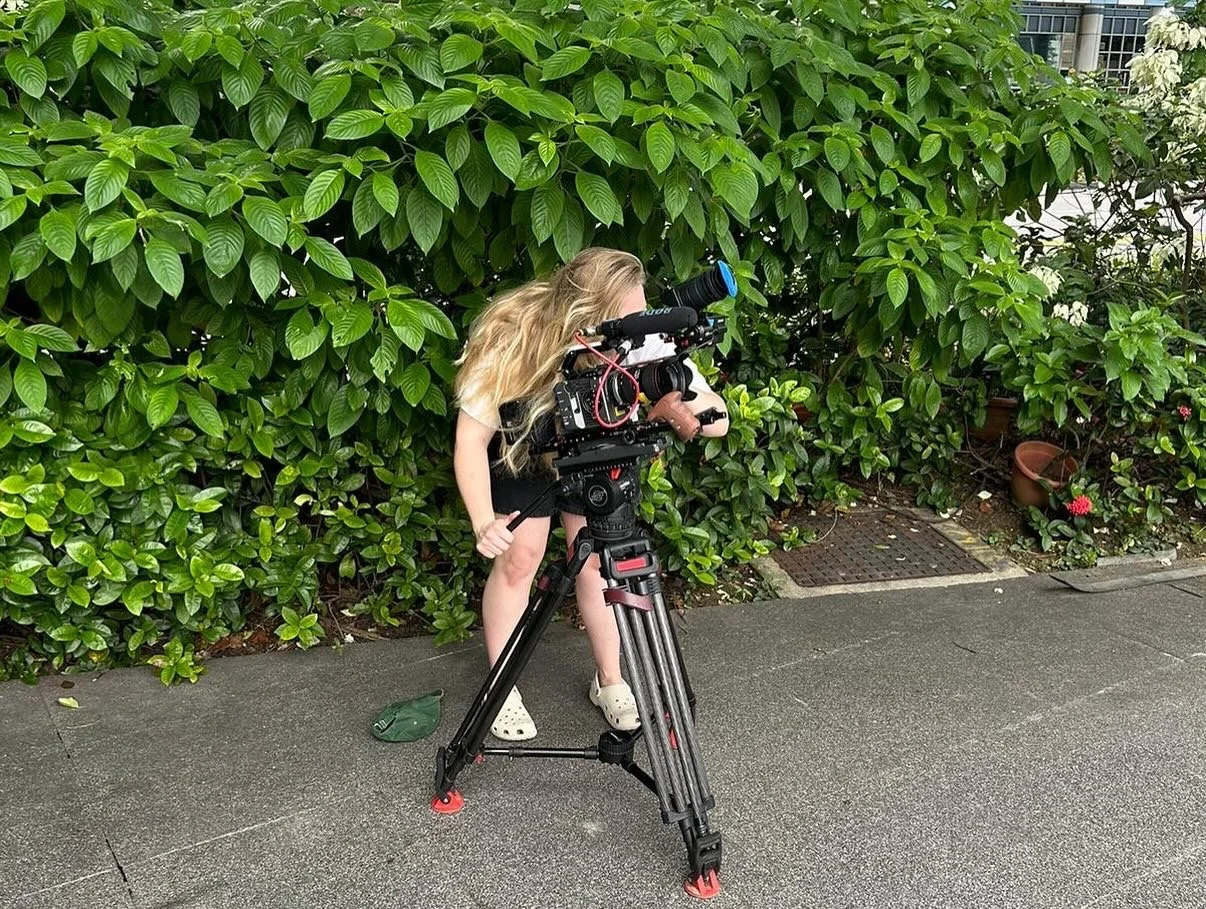 A girl operating a professional video camera on a tripod outdoors near a leafy green bush.