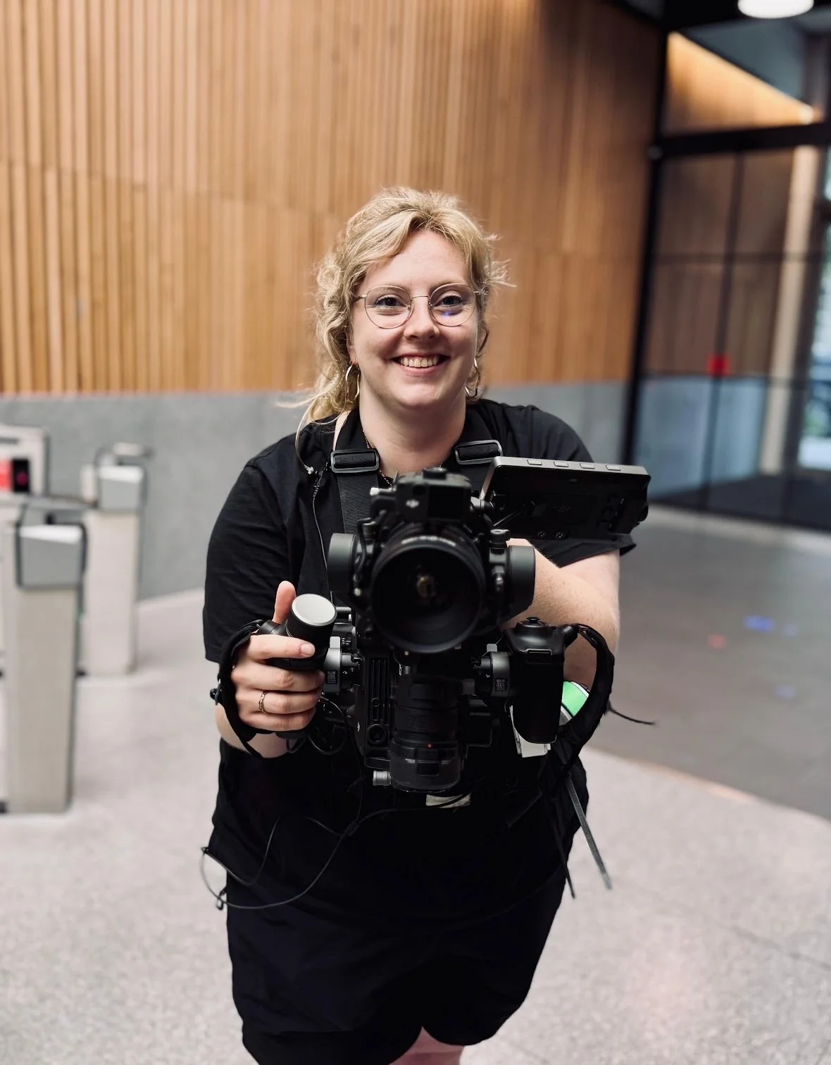 A woman with curly blonde hair and glasses smiling while holding a professional camera and monitor in an indoor setting with wooden wall panels and glass doors.