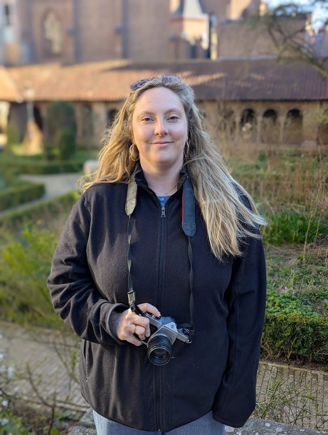 A woman standing outdoors holding a camera with a lens pointing downward. She has long blonde hair, is wearing earrings, a black zip-up jacket, and gray pants. The background features a garden with greenery and rocks, with a brick building and trees in the distance.