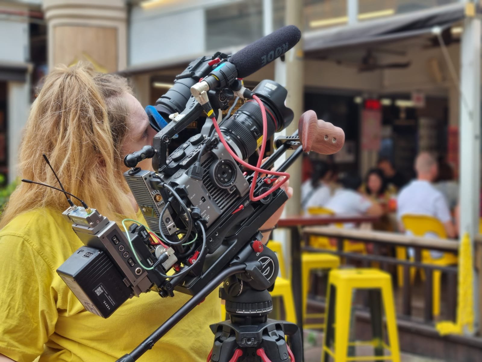 Film crew member operating a professional camera on a tripod at a restaurant or cafe with people dining in the background.