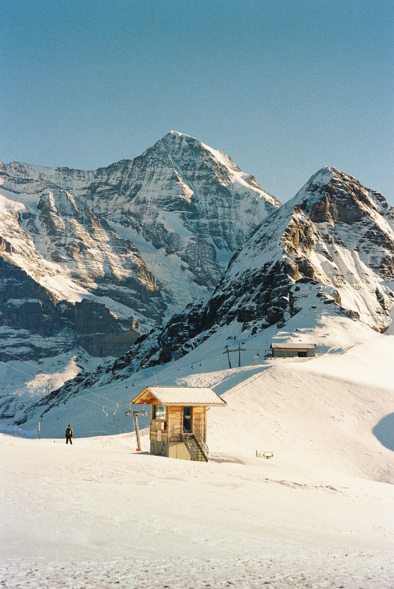 Snow-covered mountain landscape with a small wooden building in the foreground, two figures, and ski lift poles, under a clear blue sky.