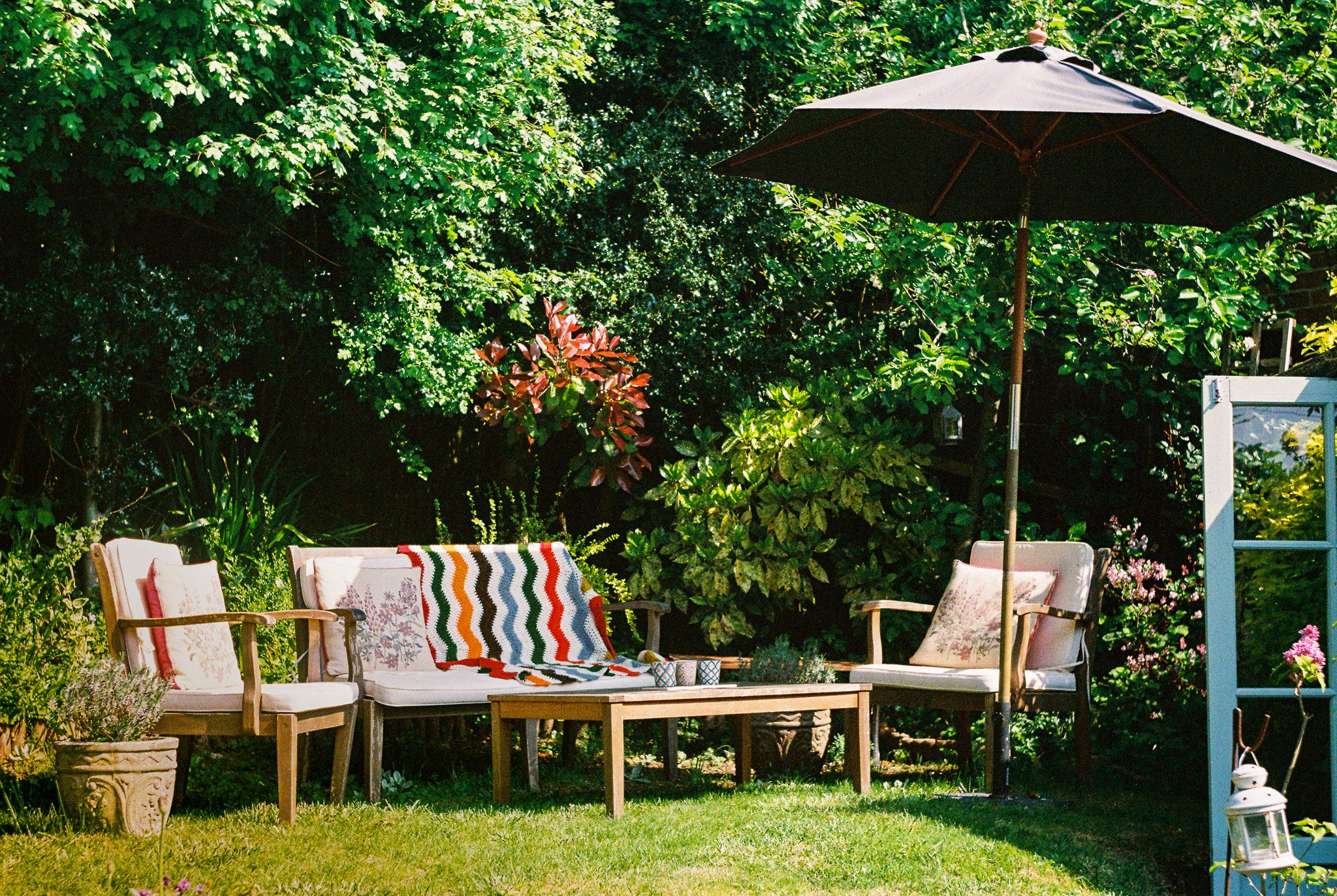 A backyard patio scene with wooden chairs, a table, a large black patio umbrella, various green plants and trees, and a colorful striped blanket on one of the chairs.
