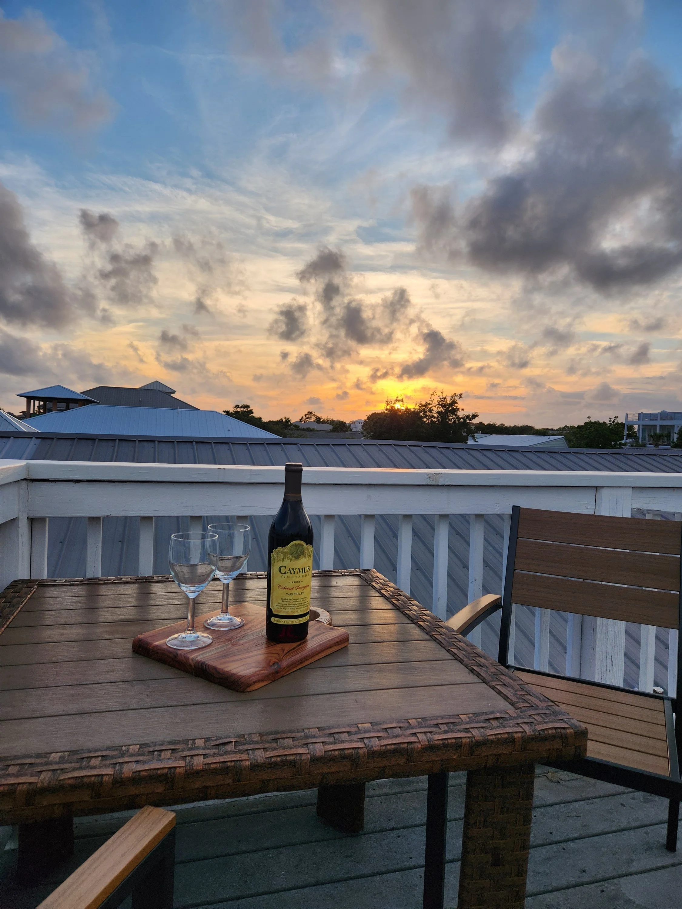 Rooftop patio table with a bottle of Caymus Cabernet Sauvignon and two wine glasses overlooking homes at sunset.