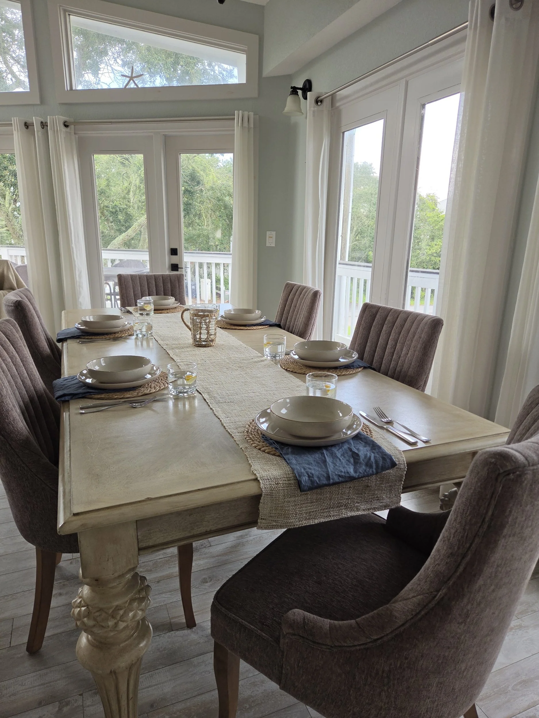 Elegant dining room with a set table, upholstered chairs, white curtains, and large windows overlooking trees.