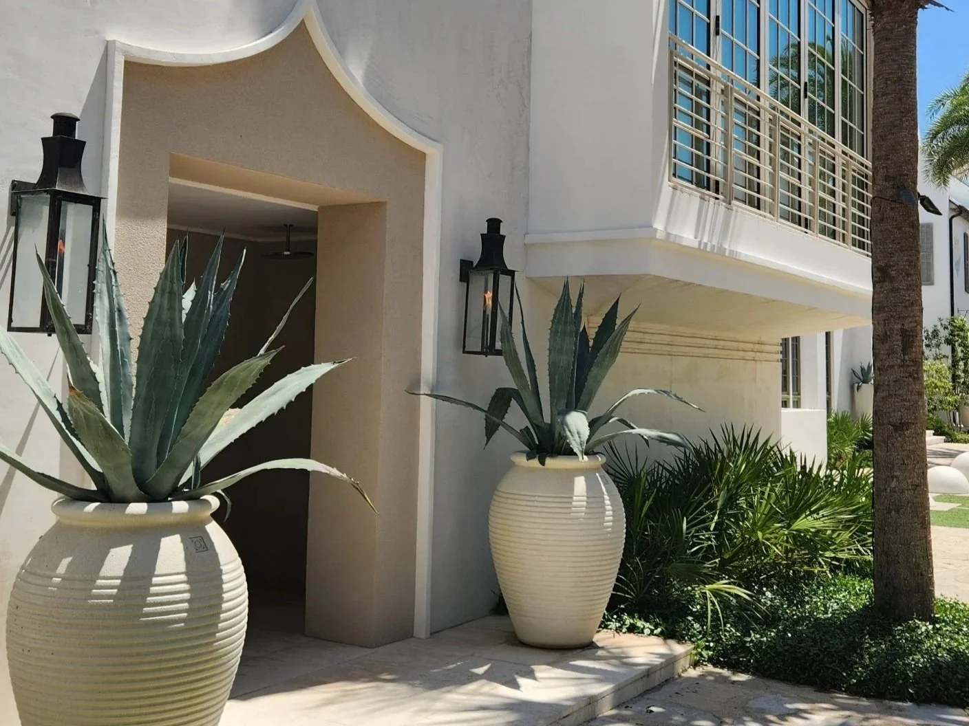 Mediterranean-style building entrance with large potted agave plants, black lanterns, palm trees, and tall gridded windows