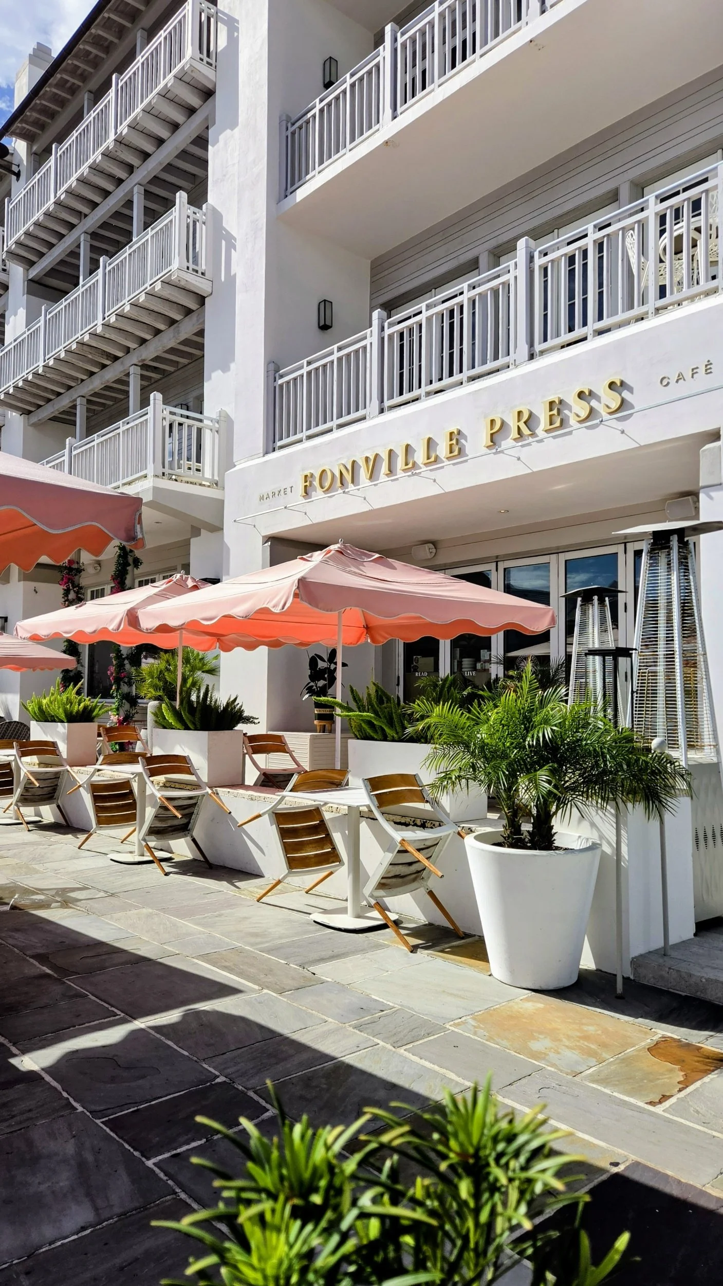 Outdoor patio at Fonville Press café with coral umbrellas, lounge chairs, tropical plants, and white balcony railings