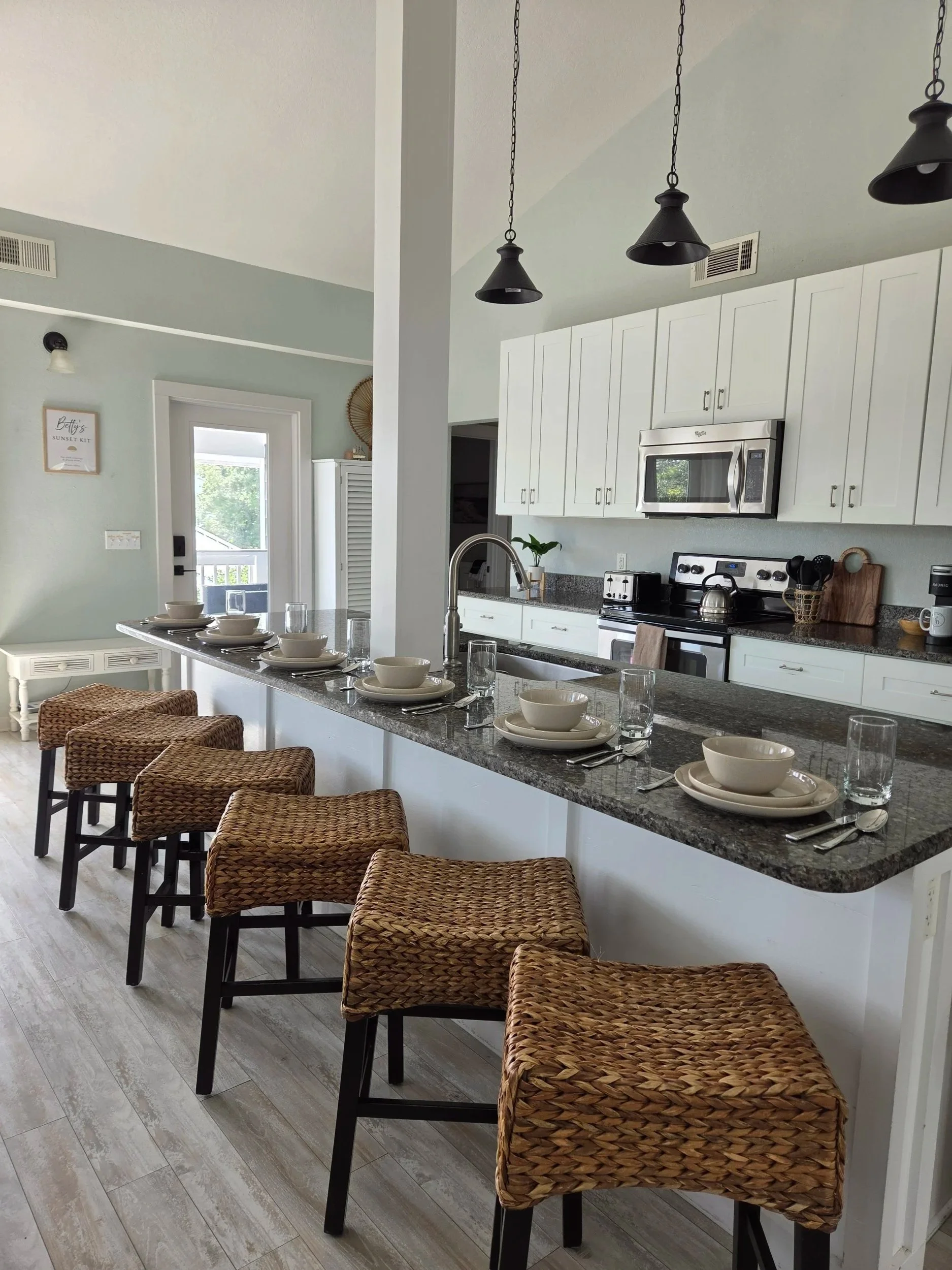 Bright home kitchen with white cabinets, granite island, wicker bar stools, pendant lights, and a fully set breakfast bar.